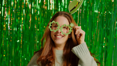 Woman-Celebrating-St-Patrick's-Day-Standing-In-Front-Of-Green-Tinsel-Curtain-Wearing-Prop-Shamrock-Shaped-Glasses